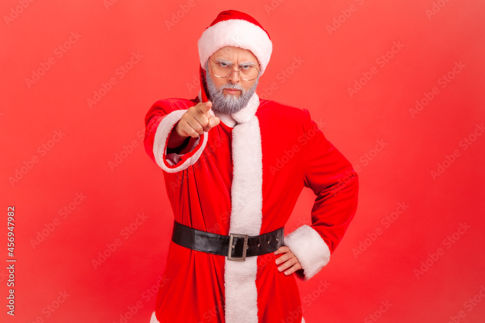 Elderly man with gray beard in santa claus costume pointing at camera with angry expression and accusing blaming, indicating at you, making choice. Indoor studio shot isolated on red background. Elderly man with gray beard in santa claus costume pointing at camera with angry expression and accusing blaming, indicating at you, making choice. Indoor studio shot isolated on red background.