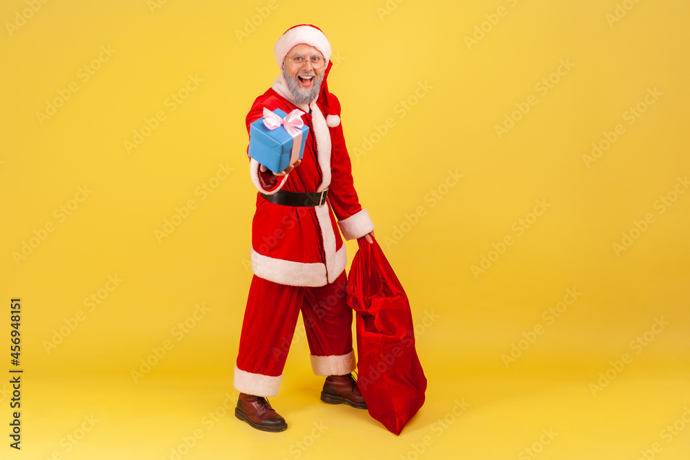 Full length portrait of elderly man with gray beard wearing santa claus costume standing with red bag with gifts and holding out Christmas present box. Indoor studio shot isolated on yellow background Full length portrait of elderly man with gray beard wearing santa claus costume standing with red bag with gifts and holding out Christmas present box. Indoor studio shot isolated on yellow background