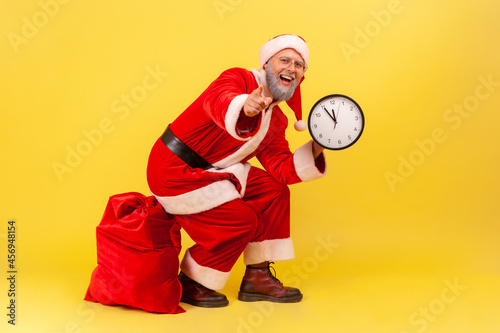 Happy positive elderly man with gray beard in santa claus costume sitting on red bag with presents, holding wall clock in hands and pointing to camera. Indoor studio shot isolated on yellow background