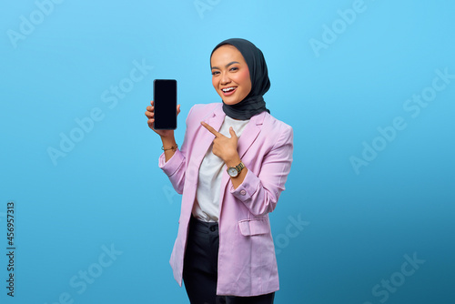 Portrait of smiling Asian woman showing smartphone blank screen over blue background