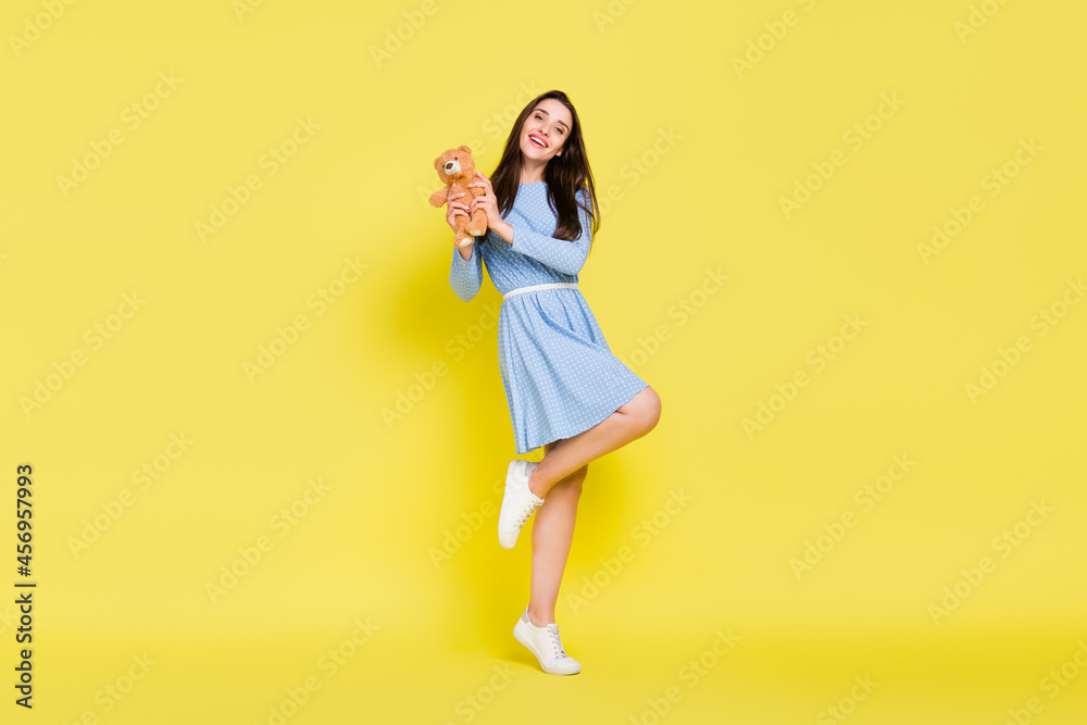 Photo of charming lady stand tiptoe hold fluffy soft bear toy wear dotted blue dress isolated yellow color background Photo of charming lady stand tiptoe hold fluffy soft bear toy wear dotted blue dress isolated yellow color background