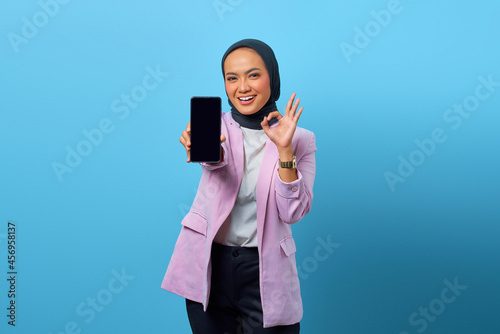 Cheerful Asian woman showing smartphone blank screen and gesturing okay sign over blue background