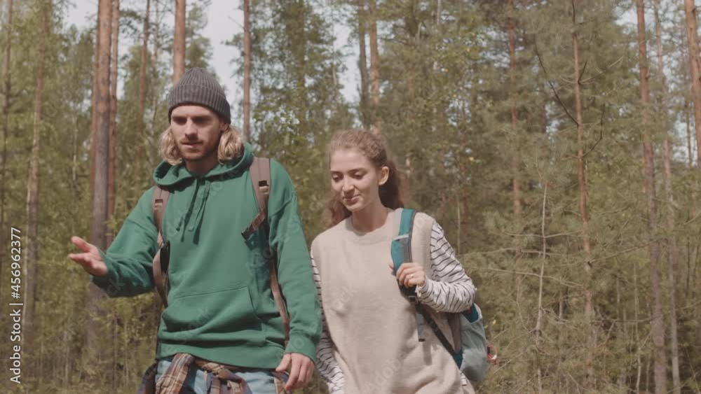 Slowmo tracking medium shot of happy young Caucasian couple with backpacks chatting while walking along beautiful summer forest Slowmo tracking medium shot of happy young Caucasian couple with backpacks chatting while walking along beautiful summer forest