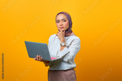 Portrait of confident Asian woman holding laptop and looking at camera over yellow background