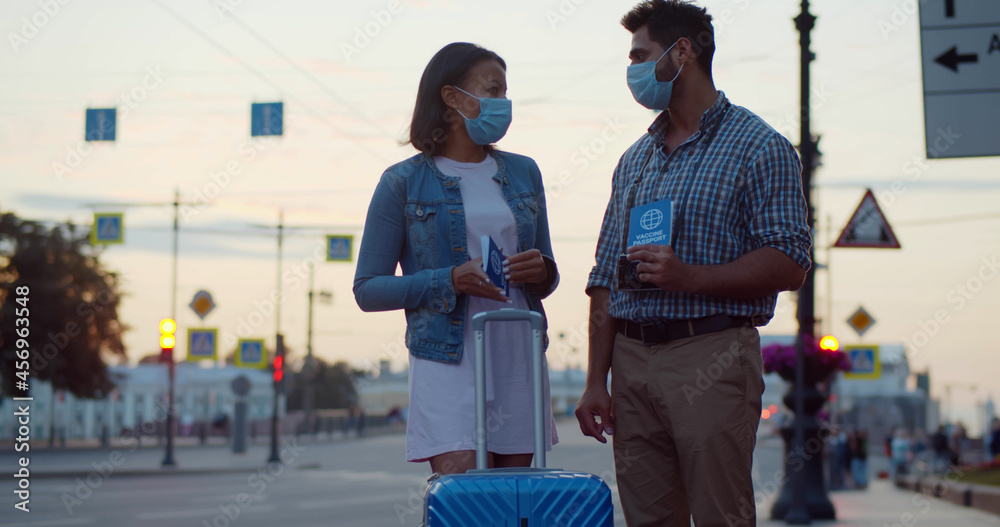 Multiethnic couple in safety mask standing outdoors with luggage and vaccine certificate