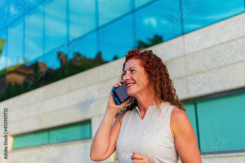 Mature 50 year old executive business woman with curly red hair and white skin with freckles smiling and talking on mobile phone outside glass office building. Horizontal view. Toledo, Spain