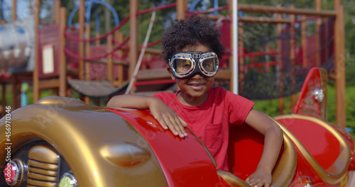 Portrait of adorable african toddler boy wearing pilot goggles sitting in toy plane on playground