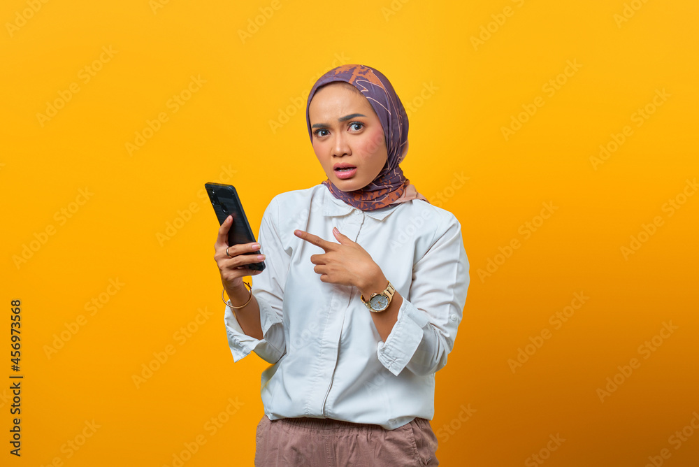 Surprised Asian woman while pointing smartphone over yellow background