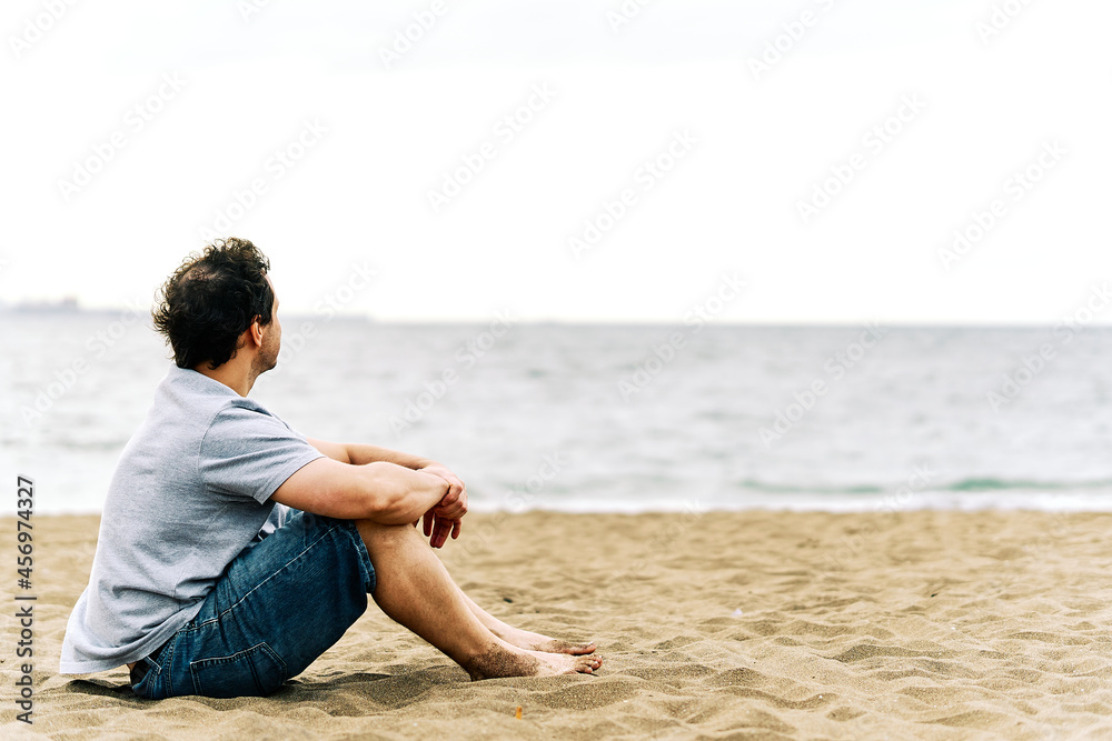 Thoughtful young adult man sitting on the beach sand with melancholic gesture