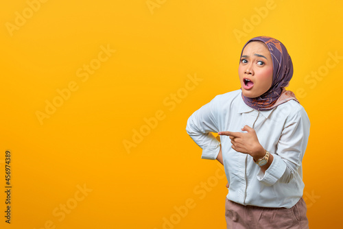 Shocked Asian woman pointing at blank space with finger over yellow background