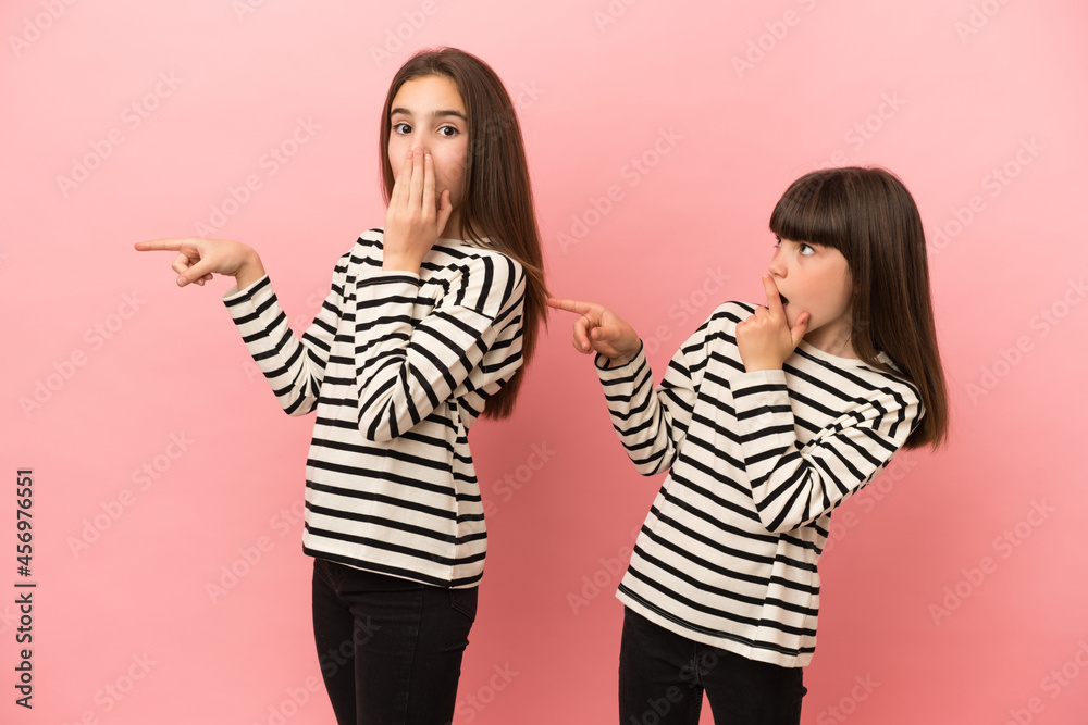 Little sisters girls isolated on pink background pointing finger to the side with a surprised face Little sisters girls isolated on pink background pointing finger to the side with a surprised face