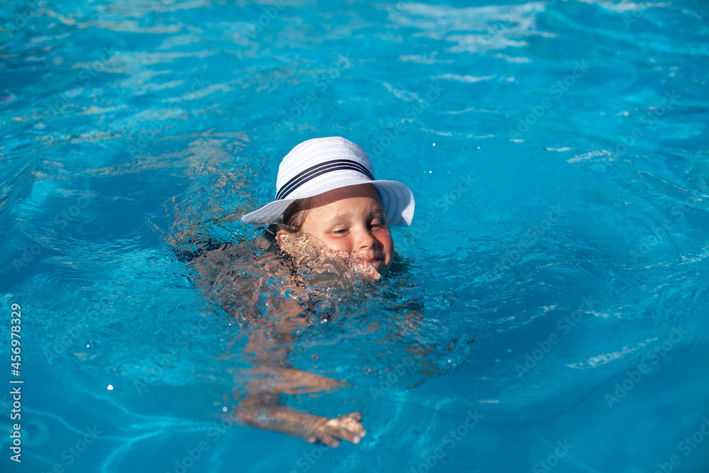 Child in white straw hat. Happy charming girl is learning swim in pool and enjoying rest on hot summer day. 