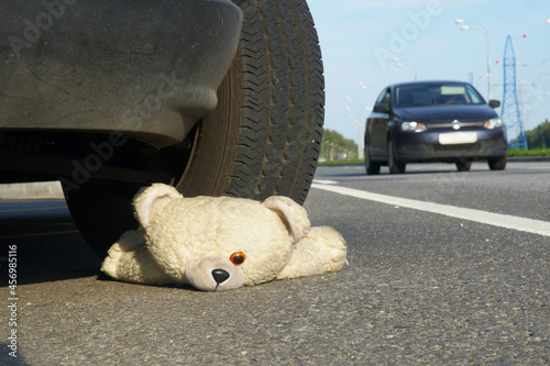 Children's toy - a teddy bear lies on the road under the wheel of a car against the background of another passing car. The concept of road accidents and road hazards.
