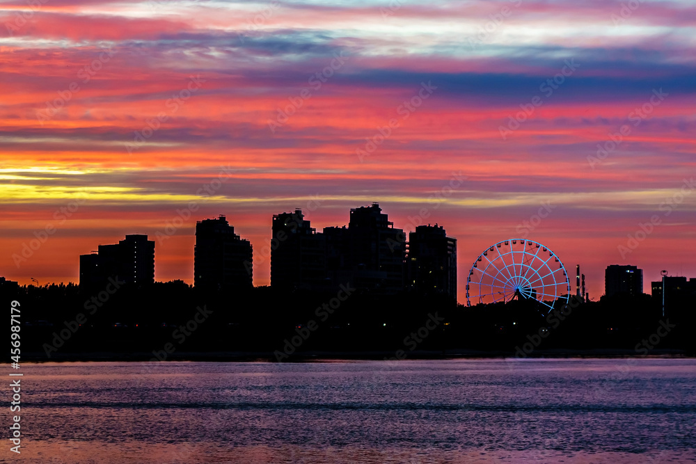Silhouette of a modern city against the background of colorful sunset clouds. There is also a river and a Ferris wheel