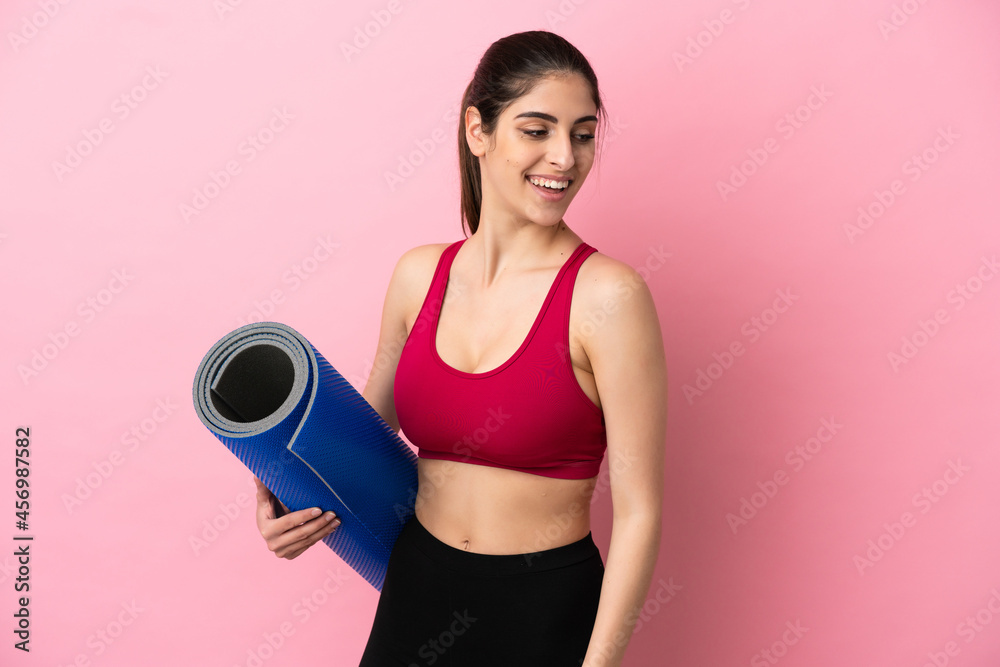 Young sport caucasian woman going to yoga classes while holding a mat looking to the side and smiling