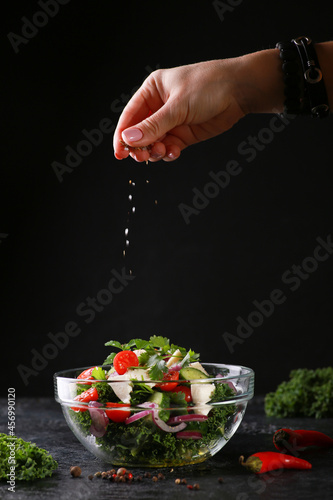 Fresh salad of herbs and vegetables in a transparent bowl on a black table. Salad of curly kale cabbage, tomatoes, cucumbers, cheese, onions and spices with hand. Background image