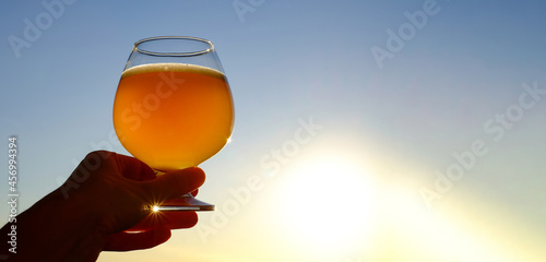 A hand holding a glass of unfiltered beer on sky background