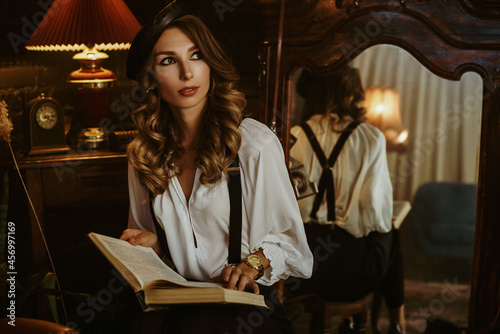 Elegant fashionable woman wearing white shirt, black beret, suspenders, trousers, holding, reading book, posing in beautiful vintage interior. Copy, empty space for text