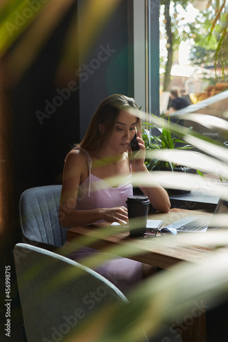 beautiful young asian woman sitting at coffee shop table surrounded by plants, working on laptop. beautiful lady calling by smartphone. modern online education, distant work, remote job, freelance