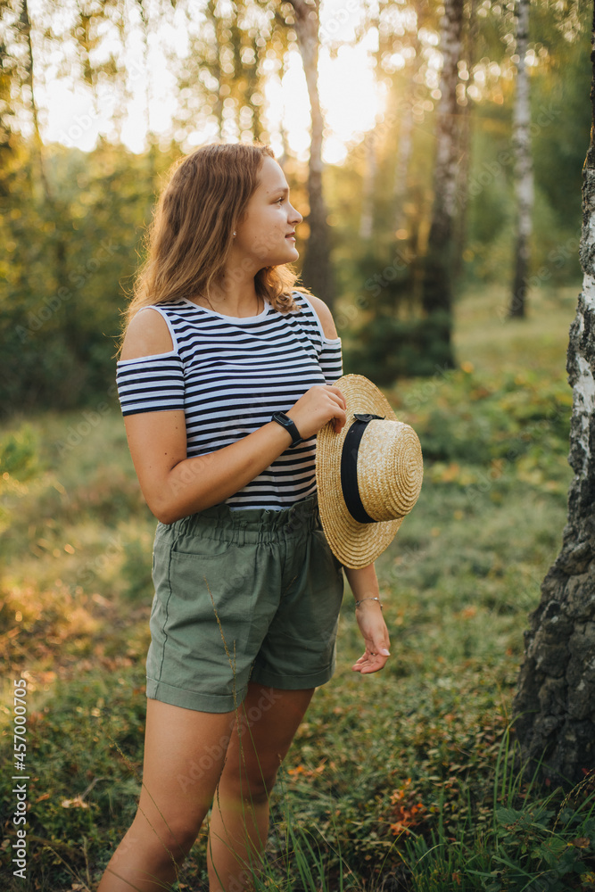 Portrait of a young girl in a straw hat. Girl in the woods at sunset. Striped t-shirt and dark green shorts