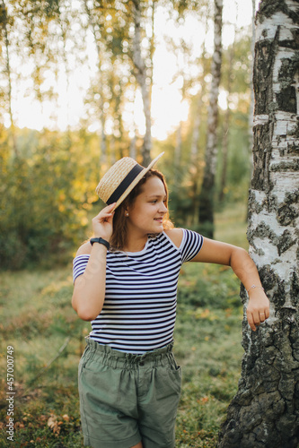 Fototapeta Naklejka Na Ścianę i Meble -  Portrait of a young girl in a straw hat. Girl in the woods at sunset. Striped t-shirt and dark green shorts
