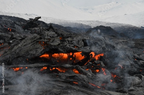 A slab of lava crust is overturned in a lava flow at Fagradalsfjall, Iceland. The lava surface is black, with red, molten lava visible beneath and steam rising. Snowy hills in the background.