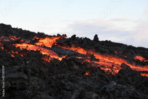Lava flow at Fagradalsfjall, Iceland. The cooled lava crust is black, while the molten lava is red and orange. Blue sky and clouds in the background.