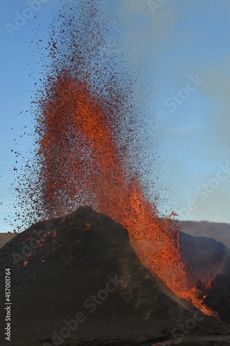 Volcanic vent at Fagradalsfjall, Iceland, erupting incandescent orange and red lava. Black cooled lava in the foreground and blue sky in the background. photo