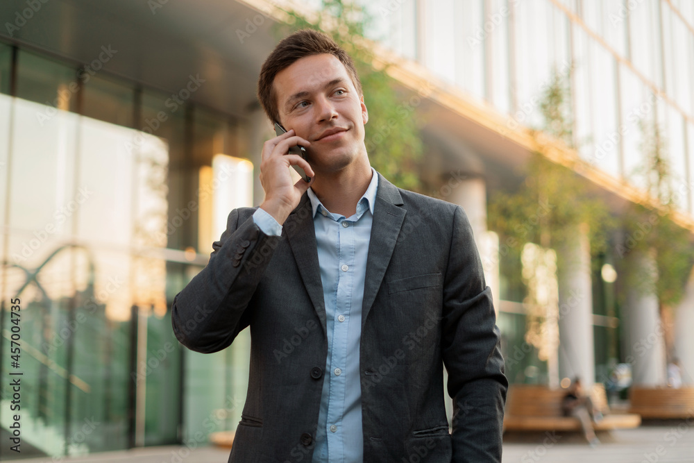 A businessman manager is calling on the phone standing near the office in a business suit. Portrait of a financier waiting for a colleague on the project. A businessman manager is calling on the phone standing near the office in a business suit. Portrait of a financier waiting for a colleague on the project.