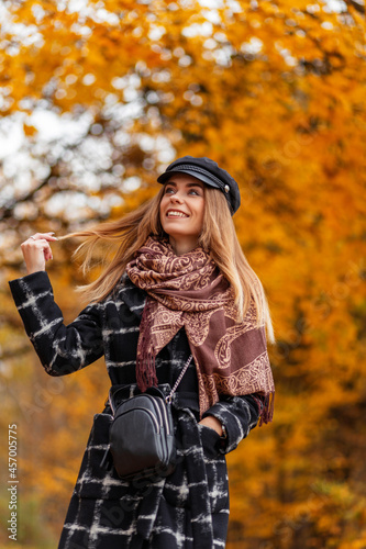 Beautiful young woman with a smile in a fashionable coat with a scarf and a hat walks in an autumn park with yellow foliage
