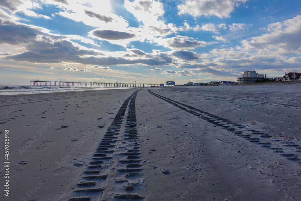 View down a deserted beach with truck tire tracks toward a long pier and a dramatic cloudscape and blue sky