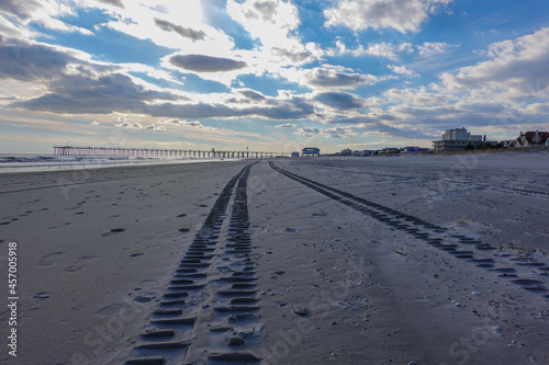 View down a deserted beach with truck tire tracks toward a long pier and a dramatic cloudscape and blue sky