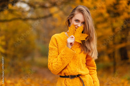 Beautiful young fashionable woman with a smile in a yellow vintage sweater covers her face with an autumn golden leaf on nature with orange fall foliage. Enjoy a happy moments