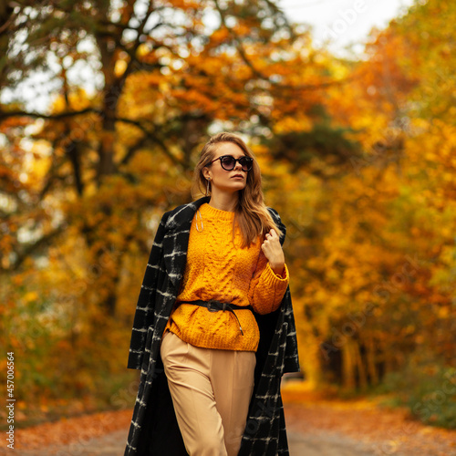 Fashionable beautiful girl with sunglasses in vintage clothes with a stylish black coat and a knitted sweater walks in the park on the nature