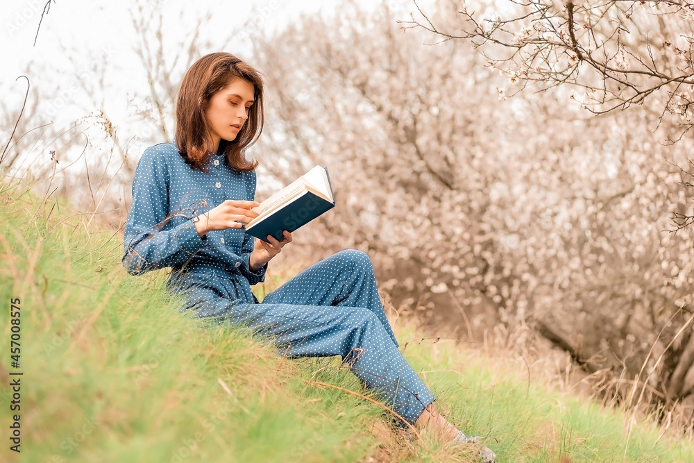 Obraz premium a girl in a blue overalls sits on the grass in the park, holds a book in her hands and reads a book, enjoying nature, preparing for exams, horizontal image.