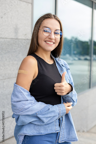 Vaccination, medicine, pandemic and healthcare concept - An excited oriental girl with dark hair and glasses with a band-aid glued on her arm after a vaccination in the street showing her thumbs up