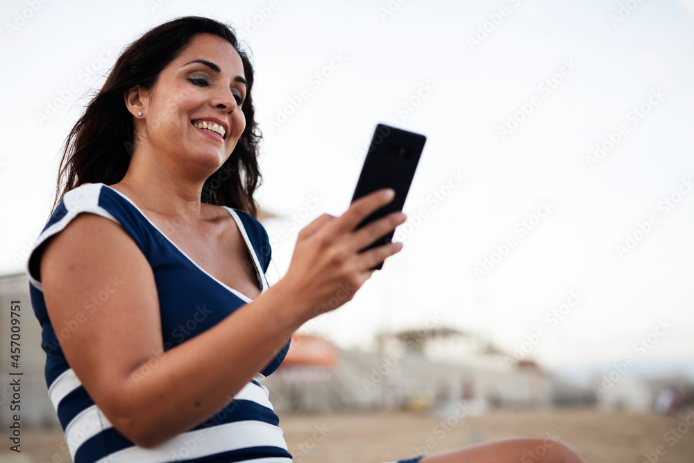 Beautiful young woman using the phone at the beach. Smiling happy woman typing a message while resting at the beach.. Beautiful young woman using the phone at the beach. Smiling happy woman typing a message while resting at the beach..