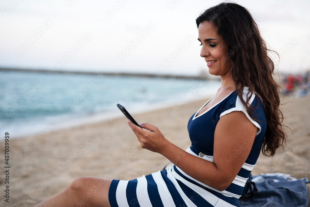 Beautiful young woman using the phone at the beach. Smiling happy woman typing a message while resting at the beach..