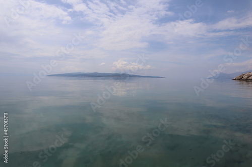 Calm at sea and clouds on a summer sunny day. Clouds reflecting on the surface of calm sea water, island on the horizon in haze, Croatian seaside