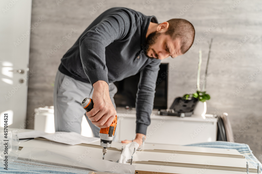 One Man with beard alone Putting Together Self Assembly Furniture at Home holding electric screwdriver screwing and putting together pieces - half length front view DIY concept real people copy space One Man with beard alone Putting Together Self Assembly Furniture at Home holding electric screwdriver screwing and putting together pieces - half length front view DIY concept real people copy space