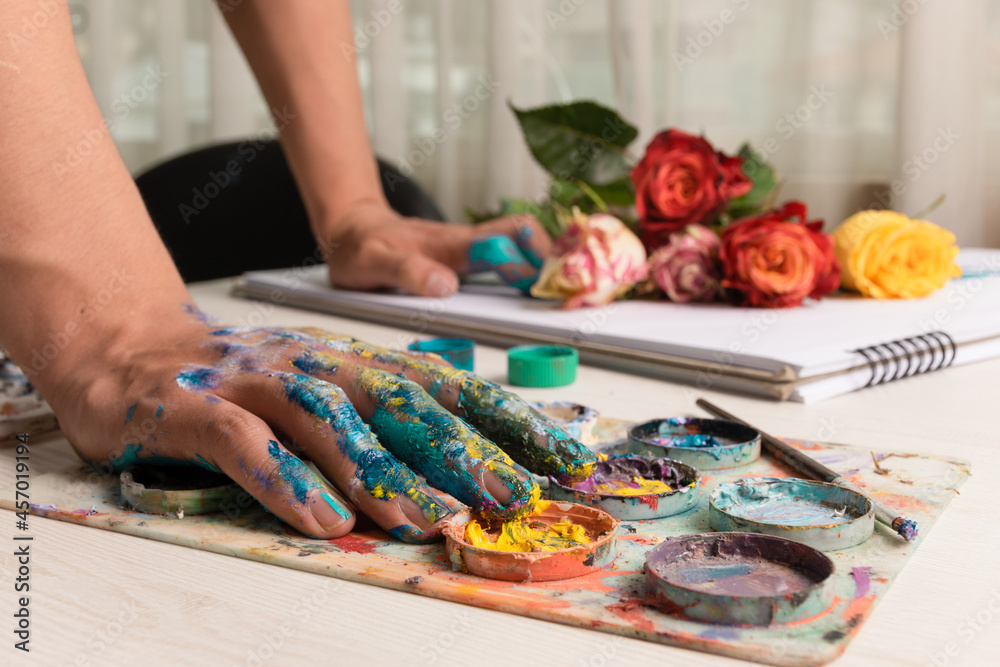 artist working with the hand of a person stained with various colors on a palette with paint on a desk with flowers and a workbook, art and creativity, lifestyle in studio