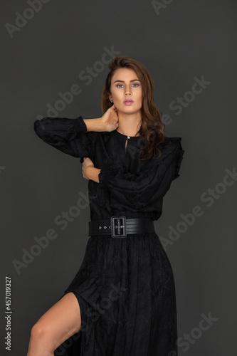 Portrait of a beautiful young white woman with wavy hair and beautiful makeup posing by herself inside a studio with a grey background wearing a black dress with a belt.