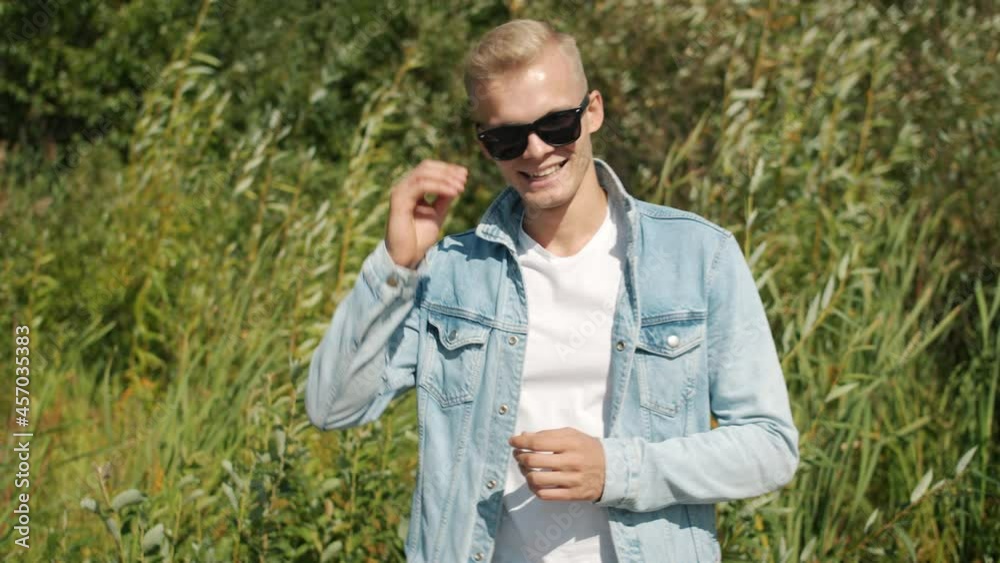 Slow motion portrait of joyful blond man standing against natural bakcground smiling looking at camera expressing joy. People and environment concept. Slow motion portrait of joyful blond man standing against natural bakcground smiling looking at camera expressing joy. People and environment concept.