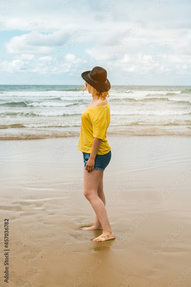 Vertical shot of a woman in a yellow shirt and black hat standing on the beach looking at the horizon with cloudy sky the beach looking out to sea under a cloudy sky