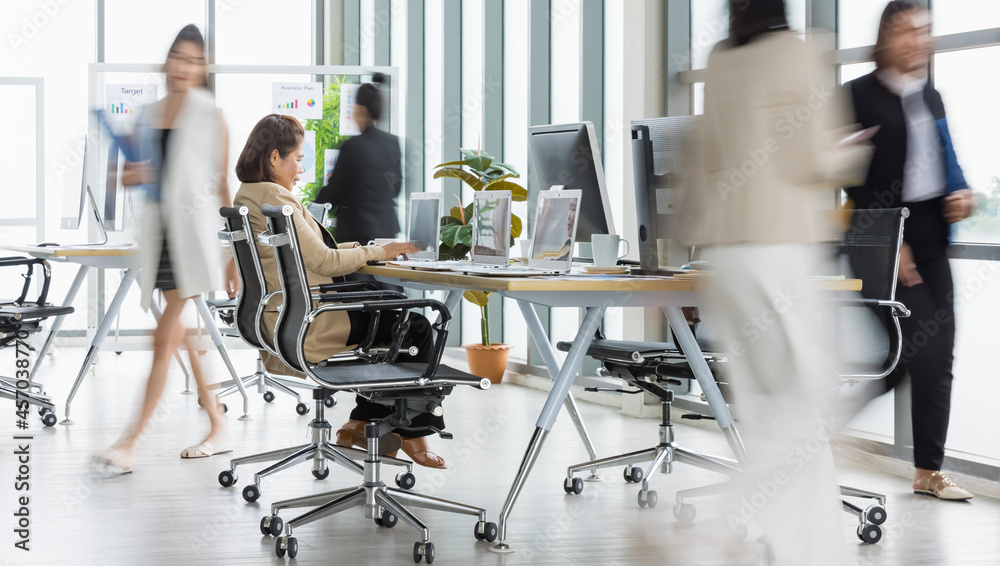 Selective focus on one business woman sitting and working on laptops on conference table with other blurry business women walking in rush in office with multiple glass windows