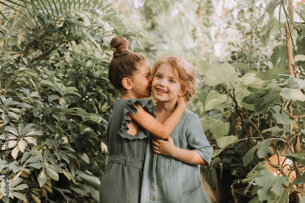 Two beautiful little girls of different nationalities walk in the botanical garden surrounded by tropical leaves and keep secrets. High quality photo