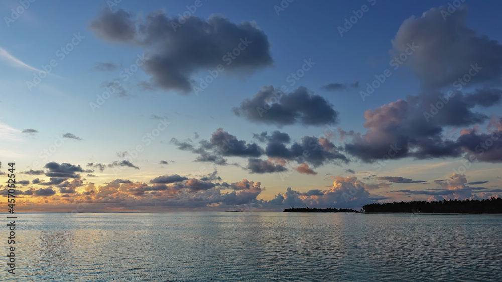 Sunrise in the Maldives. There are picturesque blue and pink clouds in the azure morning sky. Reflection and ripples on the ocean surface. An island is visible on the horizon