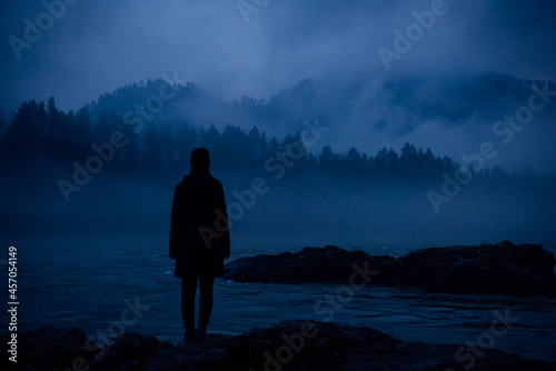 Human silhouette in a thick blue fog on the background of wooded hills and mountain river. Lonely female figure on a mysterious shore. Contemplation, meditation, unity in nature.