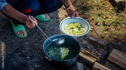 Hands of adult woman pouring soup from a cauldron into a plate with ladle. Cooking in the fresh air on a hiking trip. Large pot standing on the grass and firewood. Concept of tourism.