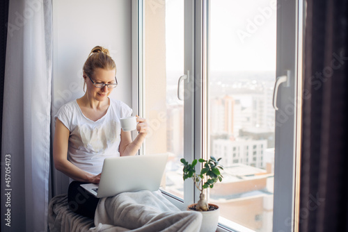 Young blonde woman wearing glasses in white t-shirt working on laptop at home, sitting on the windowsill. Freelancer and remote business. Window background. Copy space..